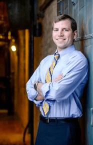 Portrait of attorney, Nathaniel F. Smith, smiling at the camera as he leans against a wall with his arms crossed. He wears a dress shirt with the sleeves rolled up, a warm light glowing in the background.