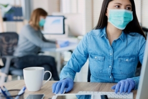 A young woman works on the computer in an office setting while wearing a blue medical mask and blue medical gloves.