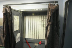 A grey voting booth, it's curtains open, ready for the next voter.